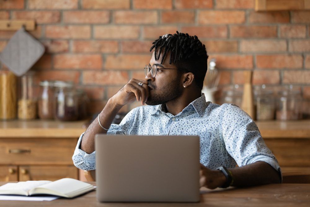 Man sits at desk with his laptop, looking away, thinking.