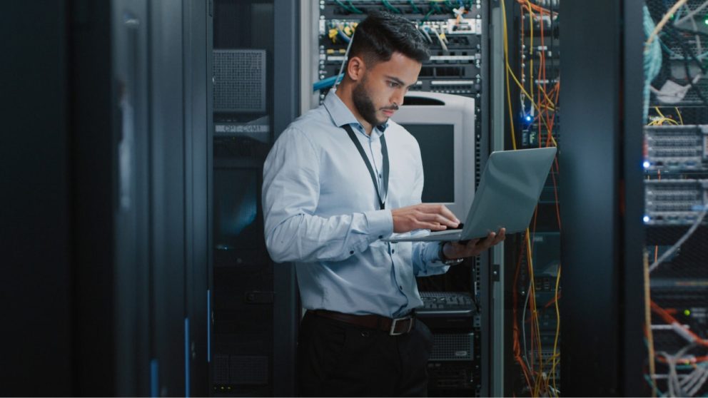 Young male IT professional working on laptop in data center server room.