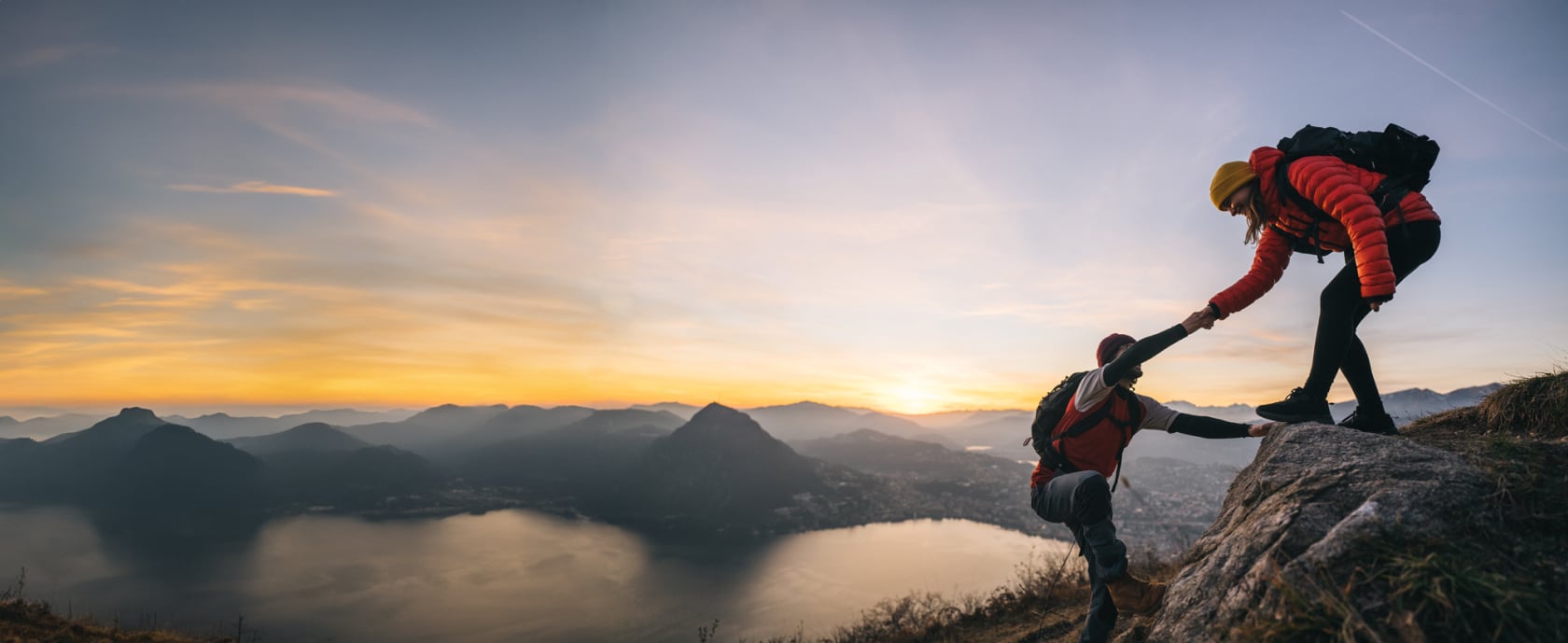 Hiker in red coat helping climbing partner reach the summit of a rock formation overlooking a valley and distant sunset.